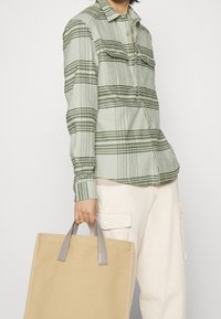 Camisa de cuadros verde claro con diseño de botones, con bolsillos en el pecho. El modelo sostiene una bolsa de mano beige con asas grises.