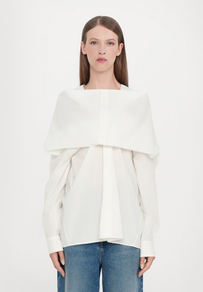 Fehér, oversized ujjas, gombos ing, amely a vállon lóg, klasszikus gallérral és egyenes aljjal rendelkezik, magas derekú kék farmernadrággal párosítva.