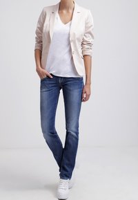 Blazer rosa claro con un solo botón, camiseta blanca de escote en V, jeans ajustados azules y zapatillas blancas. Tela suave y diseño moderno.