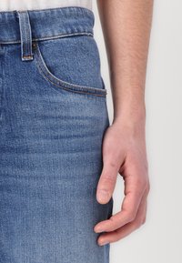 Lys blå denimjeans med middels høy midje, med et klassisk femlommers design og kontrasterende sømmer. En hånd hviler på stoffet.