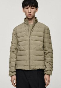 Veste matelassée en vert olive léger, avec un col haut, une fermeture zippée à l'avant et un design matelassé horizontal. Texture douce et deux poches latérales.