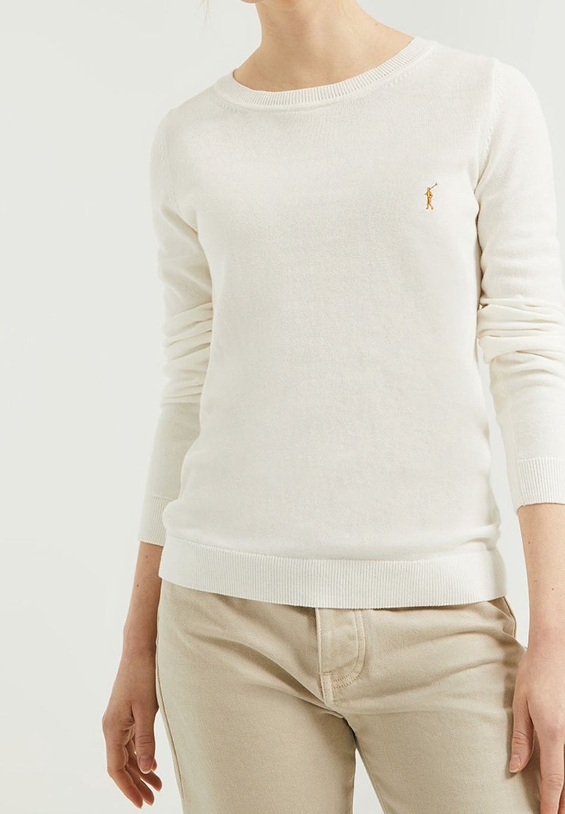 Femme portant un pull blanc à manches longues avec un petit logo brodé et un pantalon beige, montrée de la nuque aux hanches sur un fond uni.