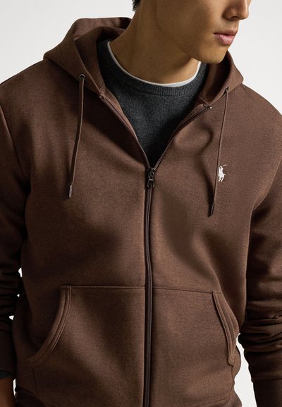 Brown zip-up hoodie with a drawstring hood, front pockets, and a small embroidered logo on the left chest. Soft texture, casual fit.