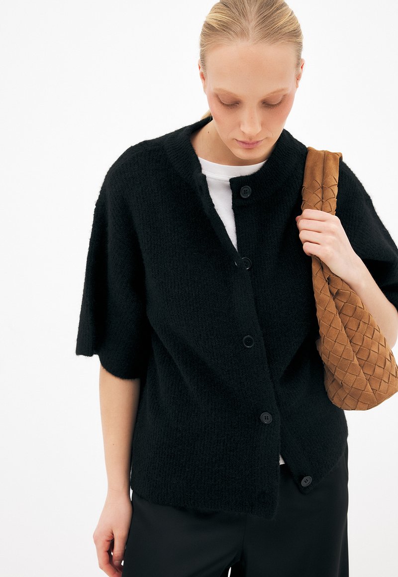 Femme portant un cardigan noir boutonné sur une chemise blanche, tenant un sac à bandoulière marron tissé, regardant vers le bas sur un fond blanc.
