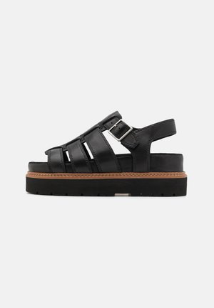 Black leather sandals with a buckle, featuring a structured design, open toe, and a thick rubber sole with a contrasting brown accent.
