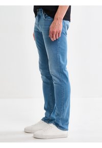 Hombre con vaqueros ajustados azul claro y zapatillas blancas, de pie contra un fondo blanco liso.