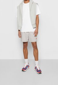 Ljusgrå ärmlös hoodie över en vit T-shirt, kombinerad med beige rutiga shorts med en orange Nike-logotyp, och flerfärgade sneakers.