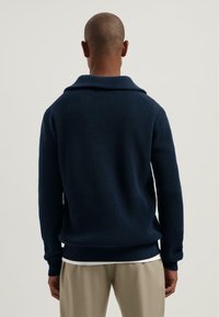 Man gezien van achteren, gekleed in een gestreepte marineblauwe trui met een brede kraag en beige broek tegen een effen lichte achtergrond.