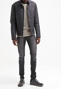 Czarna pikowana kurtka z zamkiem, jasnoszara sweter, i ciemne dżinsy. Brązowe buty na sznurowadła dopełniają strój.