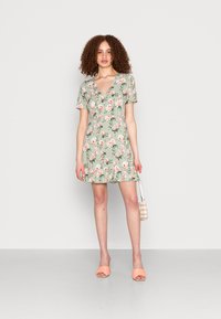 Vestido floral verde com mangas curtas, decote em V, barra com babados e rosas cor-de-rosa. Par de sandálias laranja tipo slide e pequena mala às riscas.