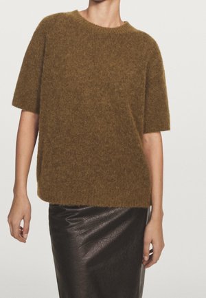 Personne portant un pull brun à manches courtes et duveteux et une jupe en cuir noire, debout devant un fond clair uni.