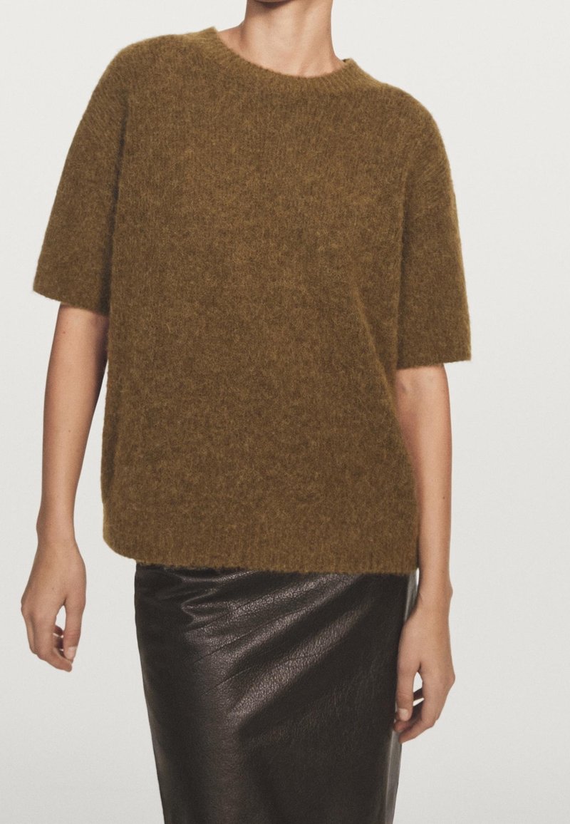Personne portant un pull brun à manches courtes et duveteux et une jupe en cuir noire, debout devant un fond clair uni.