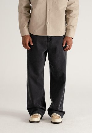 Helles beige Hemd mit Knopfleiste und zwei Brusttaschen. Schwarze weit geschnittene Jeans aus strukturiertem Stoff, kombiniert mit hellen Sneakers mit gewebten Details.