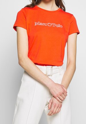 Kobieta w jaskrawo pomarańczowej koszulce Marc O’Polo i białych, wysokich spodniach z paskiem, z założonymi na sobie ramionami na wysokości talii.