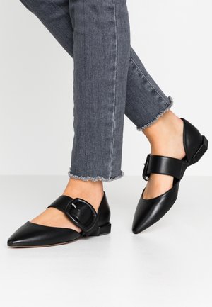 Sorte læder flade sko med spidse tæer og en ankelrem med en spænde. Båret med slidte grå jeans.