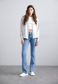 Lichtgrijze satijnen overhemd met knopen over een witte cropped top, gecombineerd met blauwe flared jeans en witte sneakers. Zachte texturen en strakke lijnen.