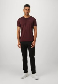 Vinrød crew-neck t-skjorte i bomull, kort erm, med en liten logo på venstre bryst. Brukes med svarte bukser og hvite joggesko.