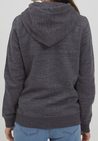 Vista trasera de una persona que lleva un sudadera con capucha gris oscuro y jeans de mezclilla azules claros, con las manos relajadas a los lados frente a un fondo simple.