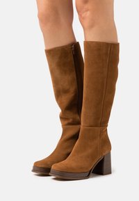 Stivali marroni in suede fino al ginocchio con tacco spesso, dotati di cerniera laterale, punta arrotondata e una texture liscia.