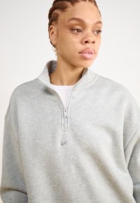 Lys grå fleecegenser med halv glidelås krage, sølv Nike-logo på brystet, og en myk tekstur, iført over en hvit topp.