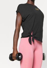 Camiseta deportiva negra de manga corta con un detalle de nudo en la cintura, combinada con leggings rosas y sosteniendo mancuernas negras. Logo de Nike en dorado.