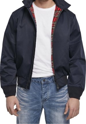 Hombre con chaqueta azul marino con forro de cuadros rojos, camiseta blanca y jeans azules desgastados, de pie con las manos relajadas a los lados.