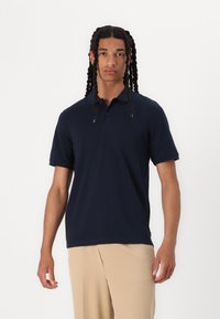Polo navy blu realizzato in morbido cotone con colletto classico, maniche corte e orlo dritto. Presenta dettagli di cucitura sottili.