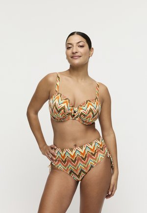 Vrouw met donker haar die een bikini met een veelkleurig zigzagpatroon draagt, poseert met één hand op haar heup tegen een effen achtergrond.