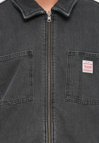 Črna denim jakna z zadrgo, dvema žepoma na prsih in strukturiranim ovratnikom. Ima vidno oznako blagovne znamke na desnem žepu.