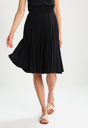 Donna che indossa una gonna nera plissettata al ginocchio, top nero e sandali beige aperti con cinturini incrociati che cammina su sfondo bianco.