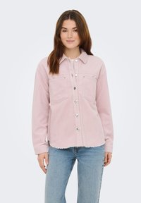 Jonge vrouw die een lichtroze corduroy jasje met knopen en lichtblauwe spijkerbroek draagt, staand tegen een effen lichte achtergrond.