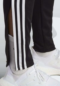 Black athletic pants feature white side stripes and zippered hems, paired with white sneakers and ribbed socks. Close-up view shows textures.