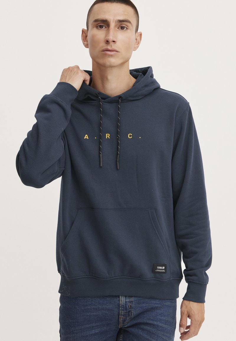 Sudadera azul marino hecha de material suave, con un bolsillo frontal, cordones y el texto amarillo "A.R.C." en el pecho. Estilo con puños acanalados.