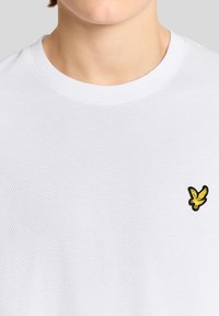 Κοντινό πλάνο ενός ατόμου που φοράει λευκό πλεκτό μπλουζάκι με στρογγυλή λαιμόκοψη και μικρό κίτρινο λογότυπο πουλιού κεντημένο στο στήθος.