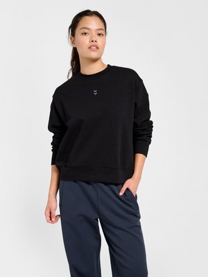 Vrouw die een zwart lange mouwen sweatshirt met een klein logo draagt en donkerblauwe broek, staand met één hand in de zak tegen een effen achtergrond.