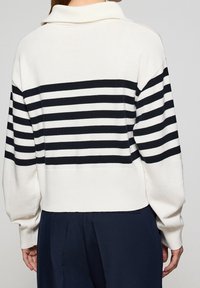 Personne portant un pull blanc à rayures horizontales noires dans le dos et sur les manches, associé à un pantalon bleu marine foncé, vue de dos.