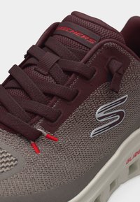 Κοντινή λήψη ενός παπουτσιού Skechers σε μπορντό και γκρι χρώμα με ύφασμα από δίχτυ, επενδεδυμένο γιακά, δετό μπροστά και παχύ λευκό πέλμα με την επιγραφή "Glide".