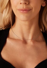 Zilveren ketting met een delicaat ontwerp, een gladde textuur en een dun profiel, rustend op een blote nek tegen een donkere stoffen achtergrond.