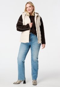 Beige opvouwbare vest met capuchon, met gestikte details op de mouwen, gecombineerd met een donkere trui en lichtblauwe jeans. Enkellaarsjes met slangenhuidprint.