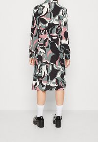 Dlouhé šaty s překříženým designem, zdobené výraznými točivými vzory v černé, růžové a tyrkysové barvě. Uvázané v pase; nošeny s robustními černými botami.