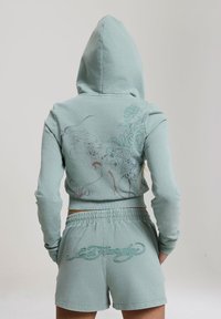 Personne portant un sweat à capuche bleu clair avec un motif floral et animal dans le dos, associé à un short assorti avec un texte brodé.