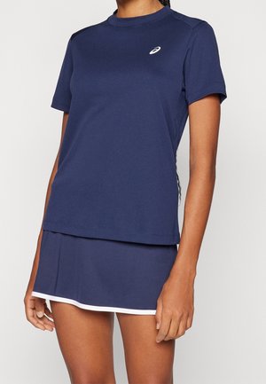 Vrouw draagt een marineblauw sportshirt met korte mouwen en een bijpassende rok met witte rand, staand tegen een effen achtergrond.