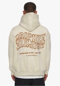 Beige hettegenser med stort, buet tekst som sier "DROPSIZE" i brunt, med «DROPSIZE CLUB / EST. 2020» under. Myk stoff, avslappet passform.
