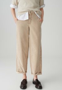 Pantaloni beige a gamba larga con vita alta, orli arrotolati e tasche laterali. Realizzati in materiale morbido con una leggera texture. Indossati con mocassini neri.