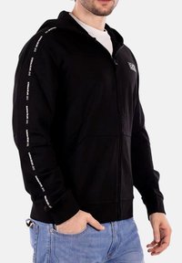 Felpa nera con zip realizzata in tessuto morbido, dotata di tasche laterali, cappuccio e testo del logo bianco lungo le maniche. Vestibilità classica.
