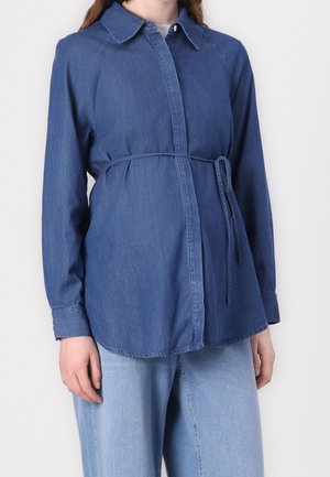 Kvinde iført en blå denim-knap-up skjorte med et tyndt bælte bundet om taljen, parret med lyseblå denimjeans.