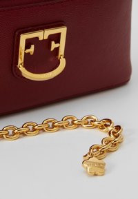 Gros plan sur un sac en cuir bordeaux avec un fermoir doré et une chaîne en or épaisse ornée d'un petit pendentif en forme de fleur gravé "FURLA".