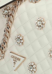Bolso acolchado en color crema con acentos de pedrería y una correa de cadena dorada. Presenta una placa de logo triangular con el texto "GUESS" y adornos.