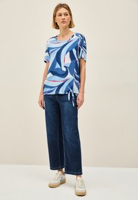 Top a maniche corte blu con disegni a spirale, dettaglio annodato all'orlo, abbinato a pantaloni in denim scuro larghi e sneaker bianche con accenti blu.