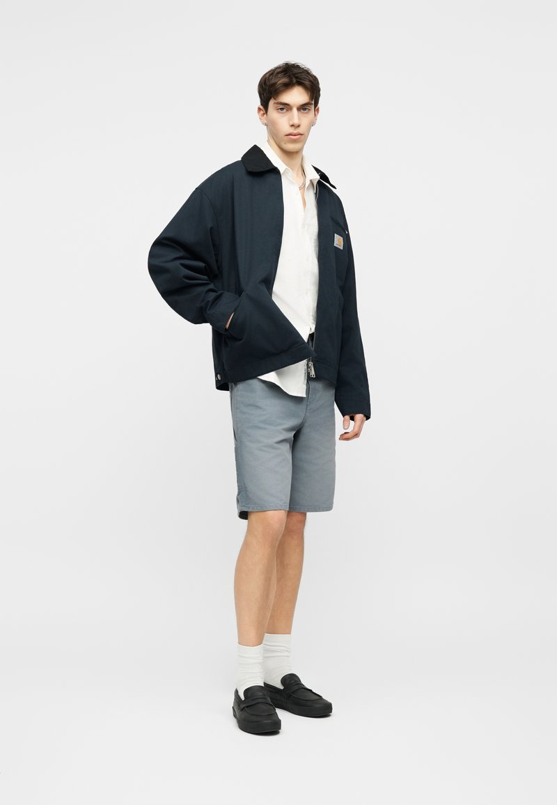 Jonge man die een zwarte jas, wit overhemd, grijze korte broek, witte sokken en zwarte loafers draagt, staand tegen een effen achtergrond.